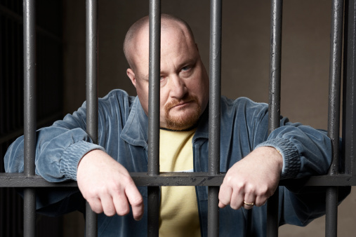 A prisoner sitting glumly behind metal bars, head lowered in frustration, symbolising the human and financial strain caused by repeated accidental releases in the UK prison system.
