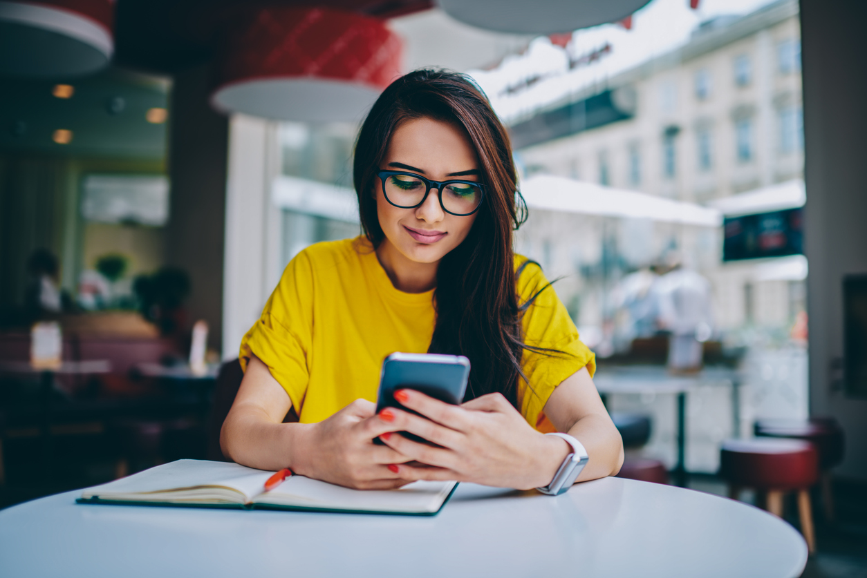 young woman blogger in spectacles publishing new post on own website and installing new application for editing photos in social networks on digital smartphone device connected to free 4g internet