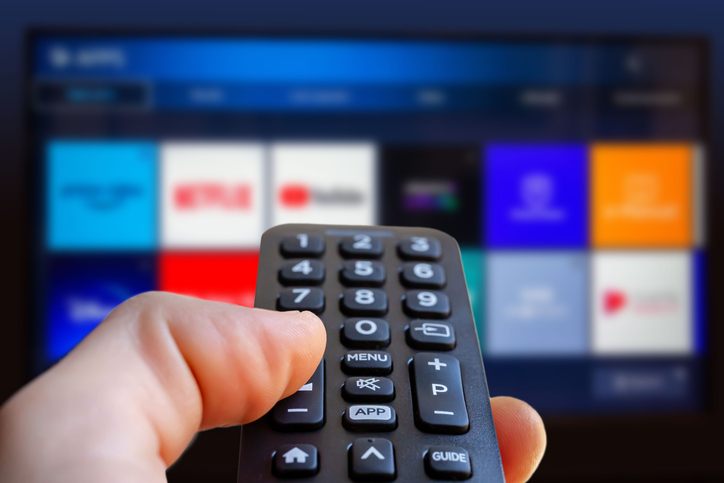 a man is holding a remote control of a smart tv in his hand. in the background you can see the television screen with streaming entertainment apps for video on demand
