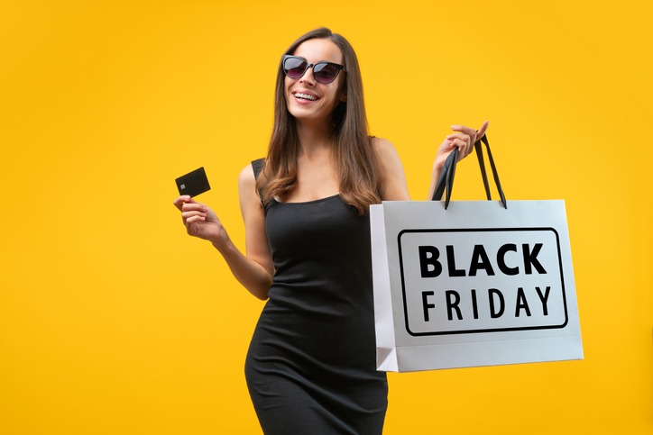 studio portrait of attractive confident young woman in trendy black dress ans glasses, posing over bright colored yellow background with credit card ans black friday shopping bag in hands