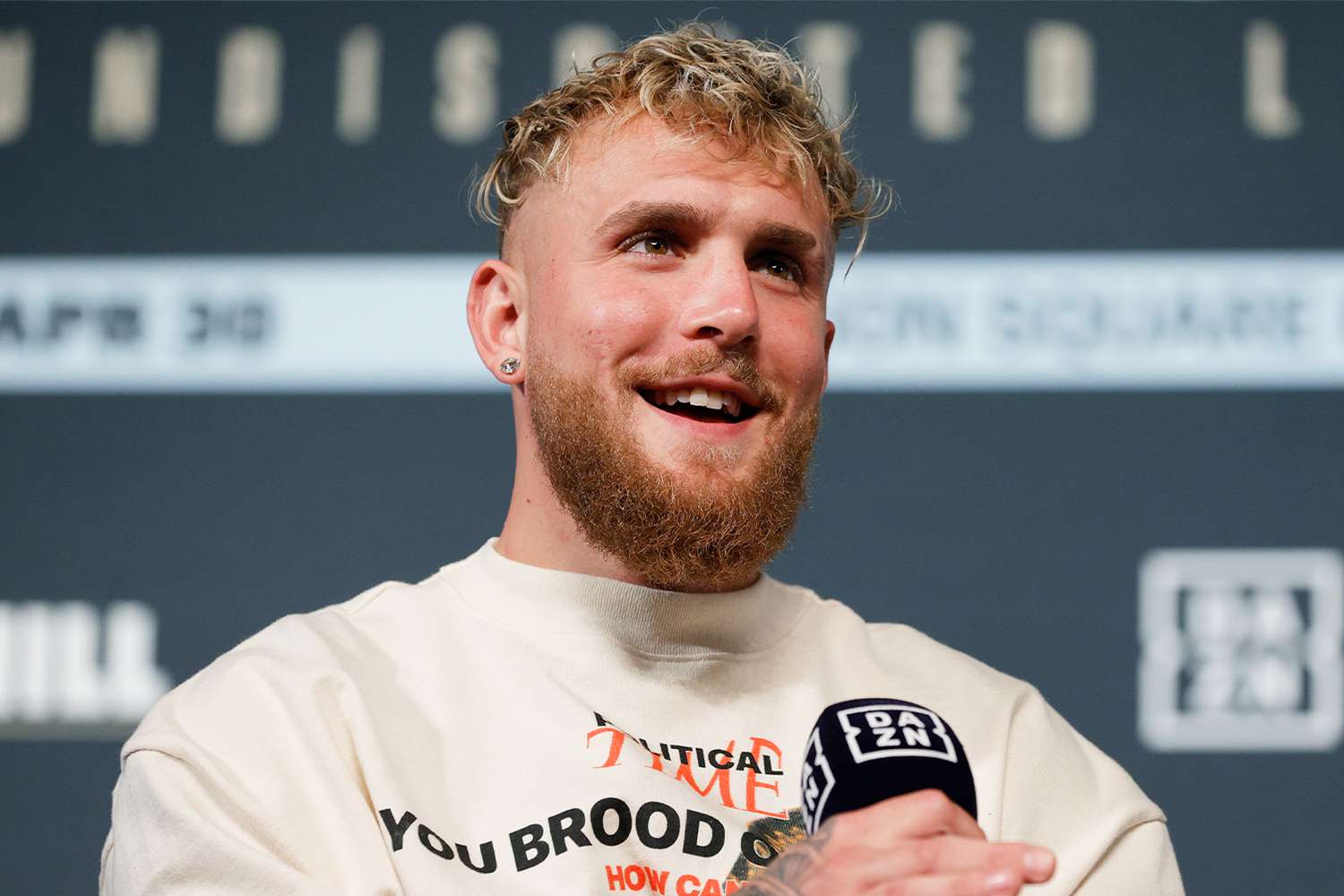 Jake Paul speaking at a DAZN press event in 2025, wearing a white sweatshirt and showcasing his $5.5 million Jacob & Co. Billionaire III watch. The boxer and YouTube star’s net worth is estimated at $100 million.