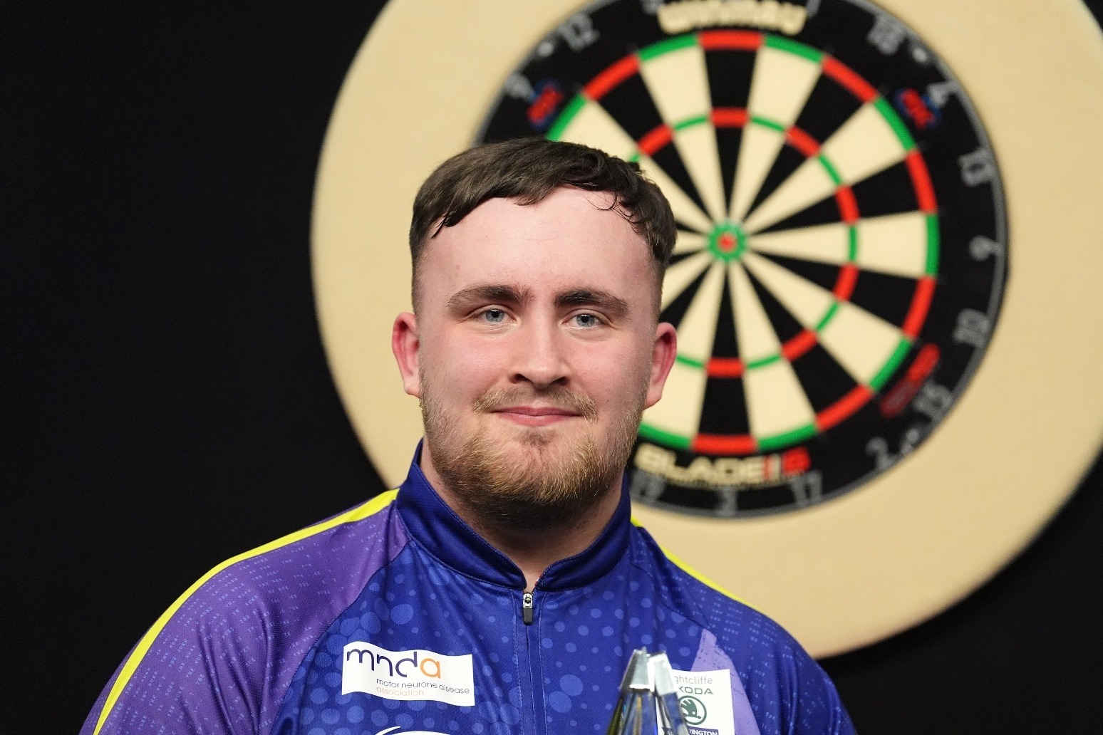 Luke Littler standing confidently in front of a dartboard, holding his darts with a focused expression.