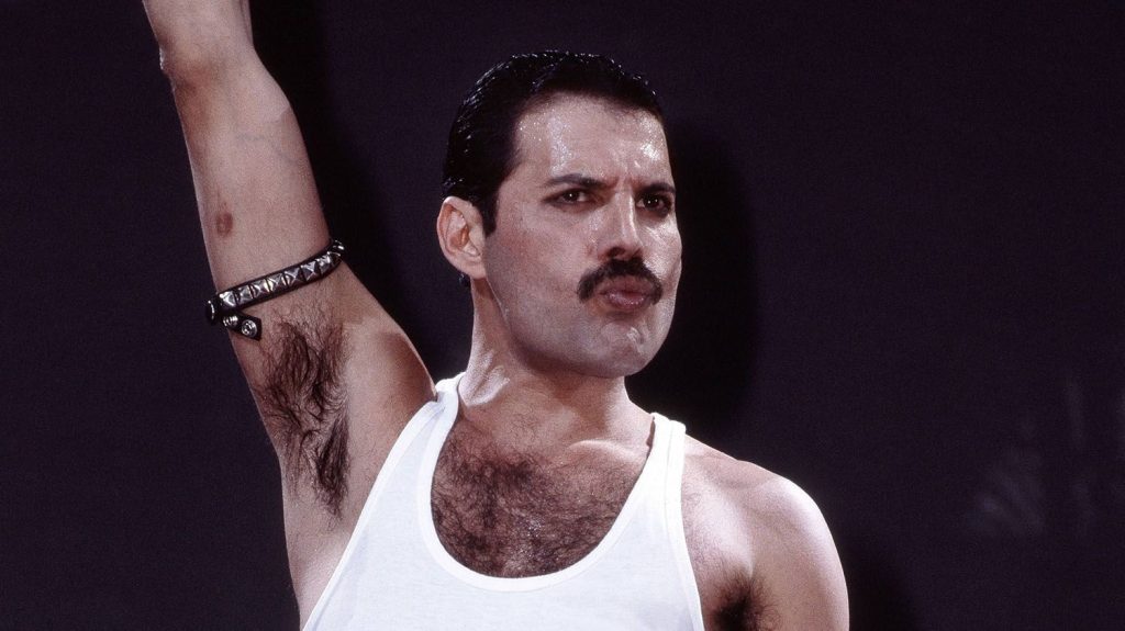 Freddie Mercury raises his arm on stage wearing a white tank top, captured during Queen's iconic Live Aid performance in 1985.