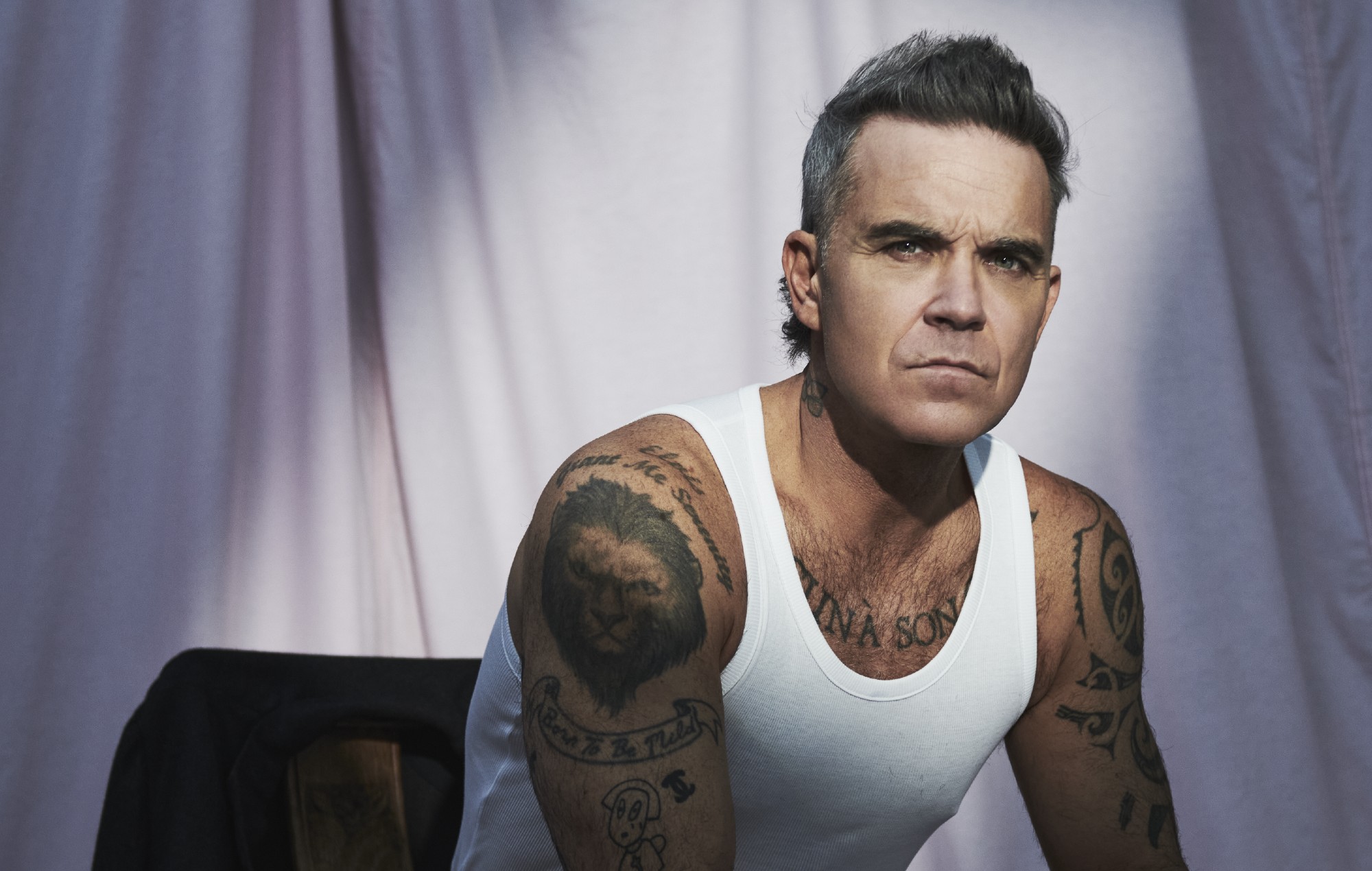 Robbie Williams posing in a white vest, looking confidently at the camera during a professional photoshoot.