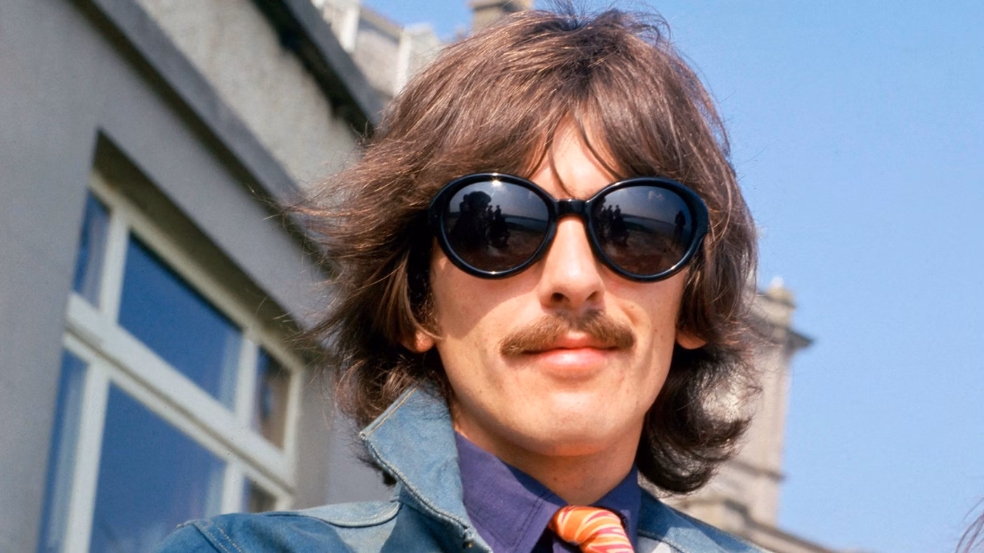 Man with long brown hair, a mustache, and oversized black sunglasses standing outside a building on a sunny day. He is wearing a blue jacket, purple shirt, and red-striped tie.