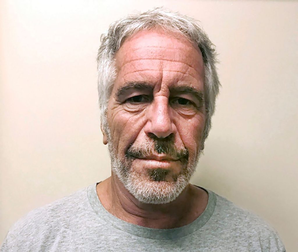 Mugshot-style photo of Jeffrey Epstein, showing him with gray hair, a short beard, and a neutral expression against a beige background.