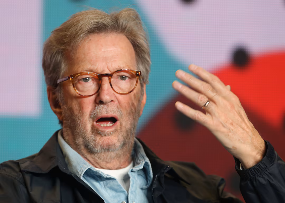 Eric Clapton speaking during an event, wearing glasses and a dark jacket, with his hand raised mid-gesture and mouth slightly open.