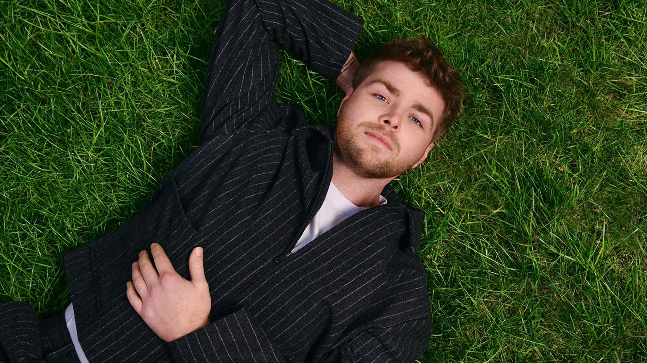 Alex Warren lying on the grass, gazing thoughtfully at the sky, dressed casually in a t-shirt and jeans.