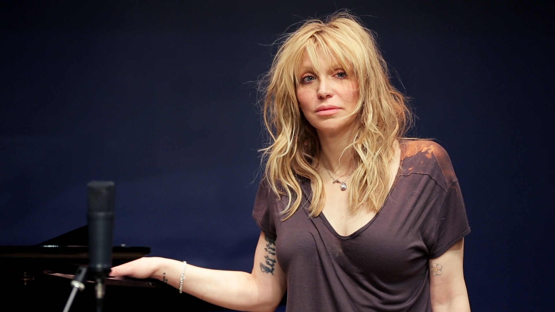 Courtney Love in a recording studio, seated at a mixing console with microphones and sound equipment around her.