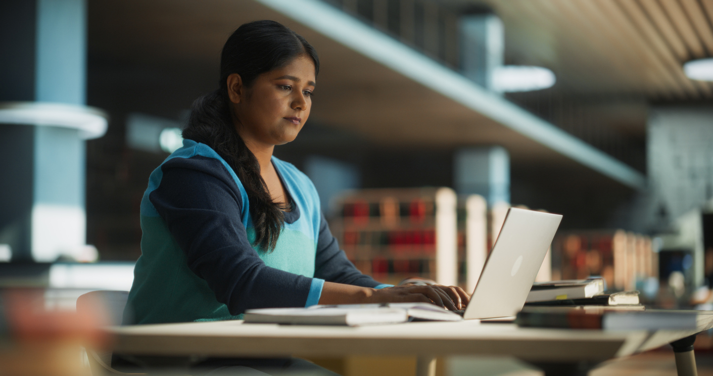 focused indian female working on a college project for class assignment, using online office software on laptop computer in a public library space. young south asian woman learning new skills online