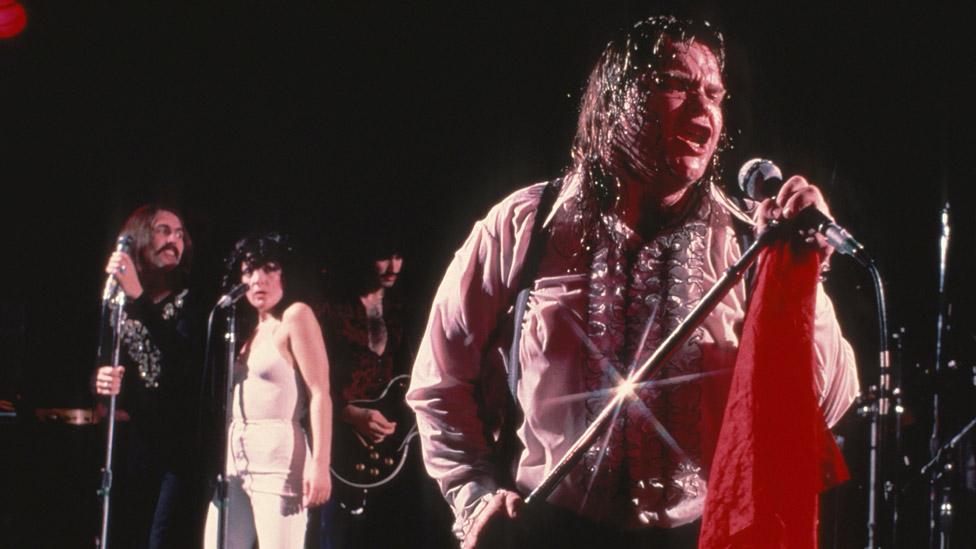 Meat Loaf blasting out powerful songs on stage with intense energy while fellow performers watch in admiration.