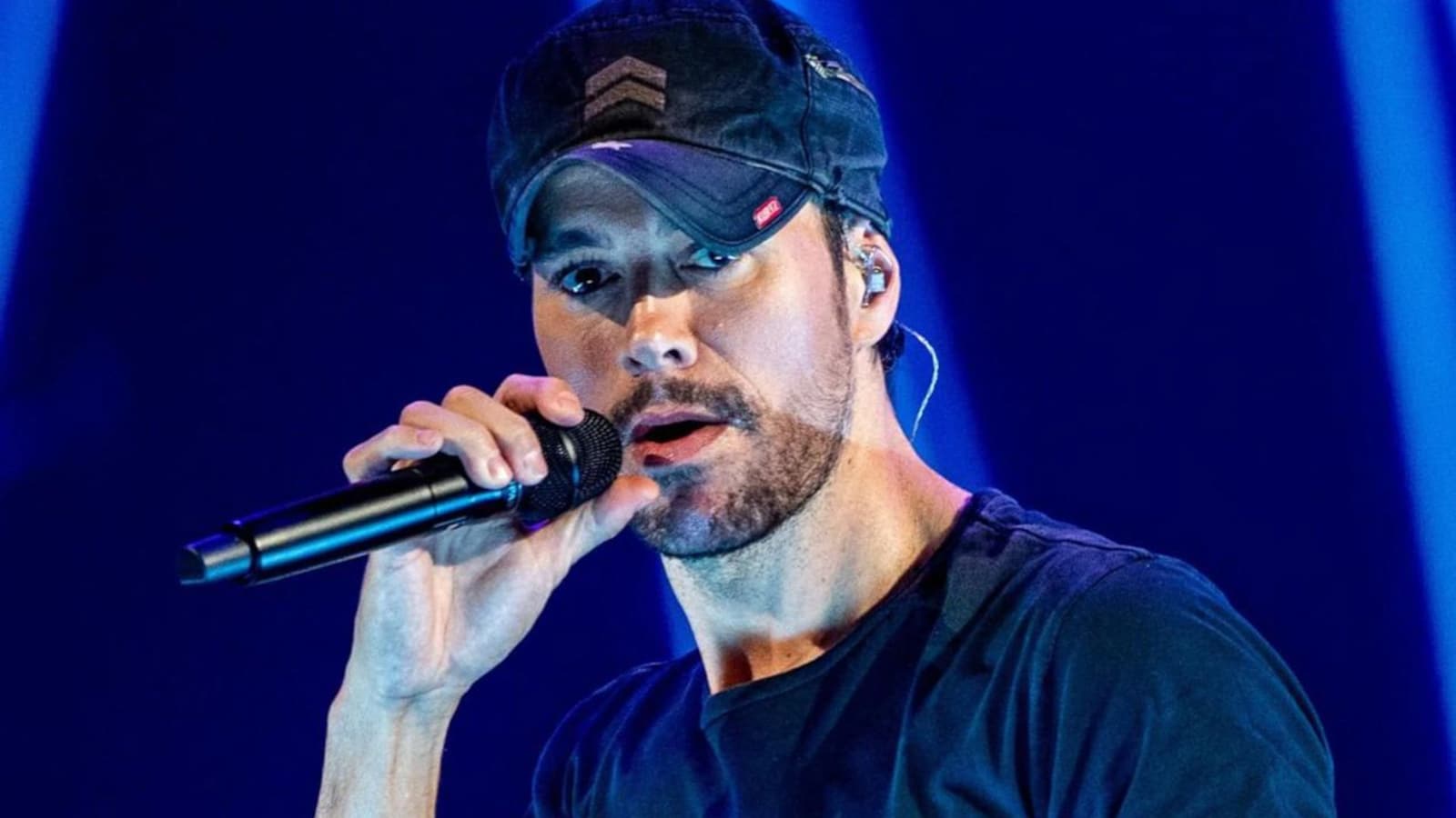 Enrique Iglesias singing on stage, wearing a black baseball cap and a dark t-shirt, passionately holding a microphone.