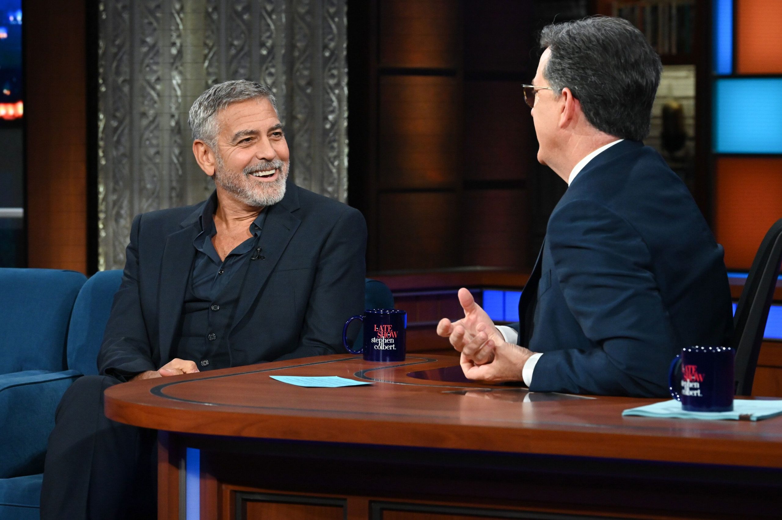 Stephen Colbert interviewing George Clooney on The Late Show set, both smiling and engaged in conversation.