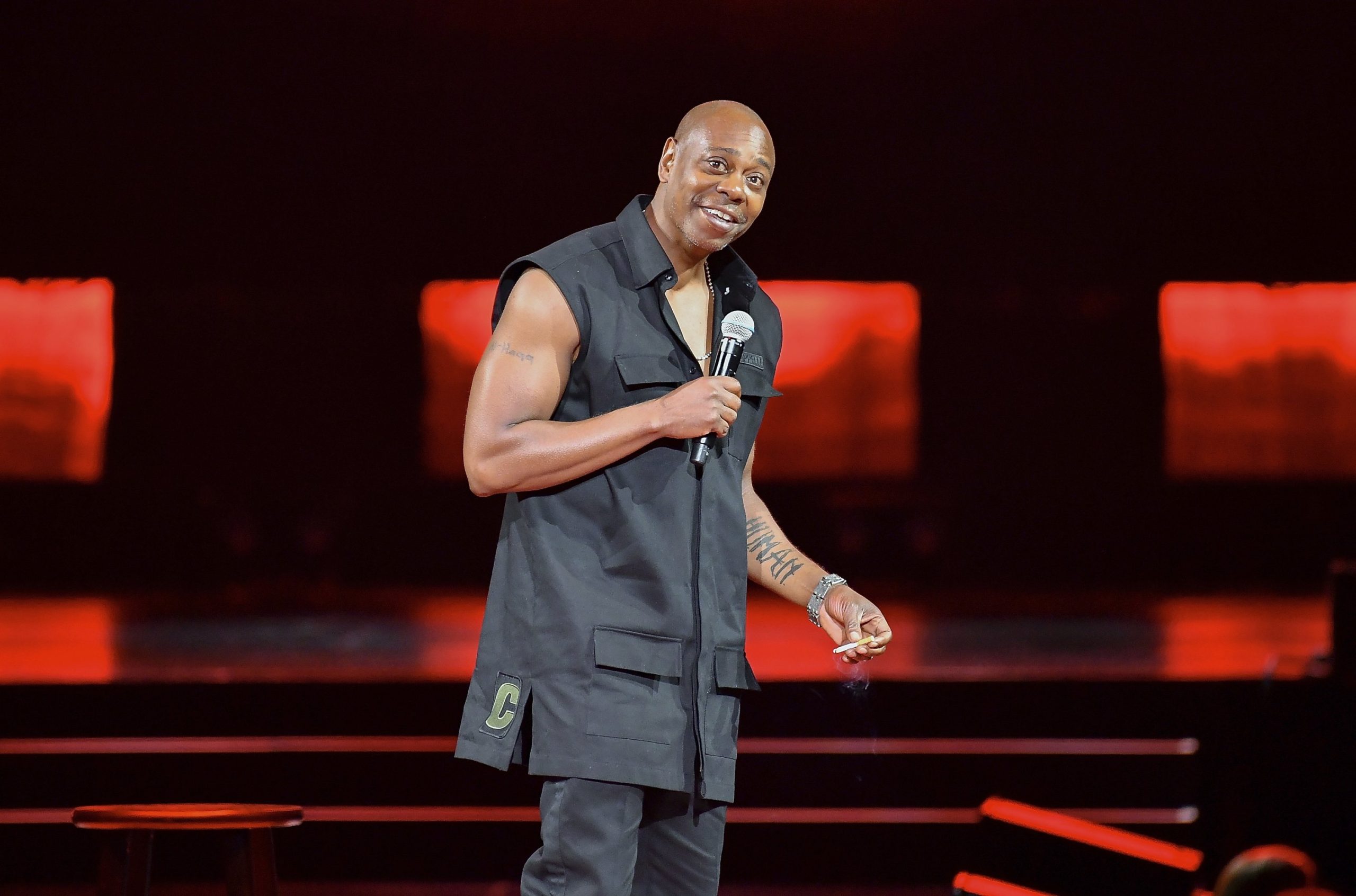 Dave Chappelle performs on stage in a distinctive sleeveless top and matching trousers, engaging the crowd with his signature style and presence.