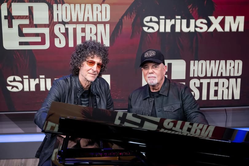 Howard Stern and Billy Joel sit side by side by a piano in the SiriusXM studio, engaged in conversation and smiling warmly.