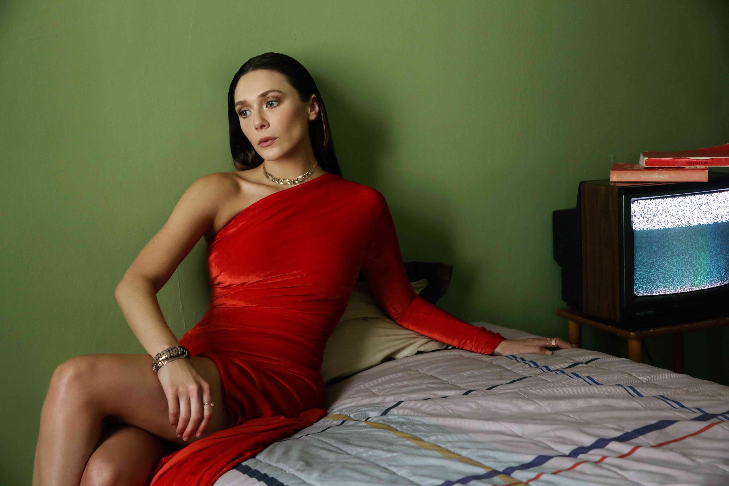 Elizabeth Olsen sits gracefully on a bed in a striking red ball gown, exuding elegance and old-Hollywood charm.