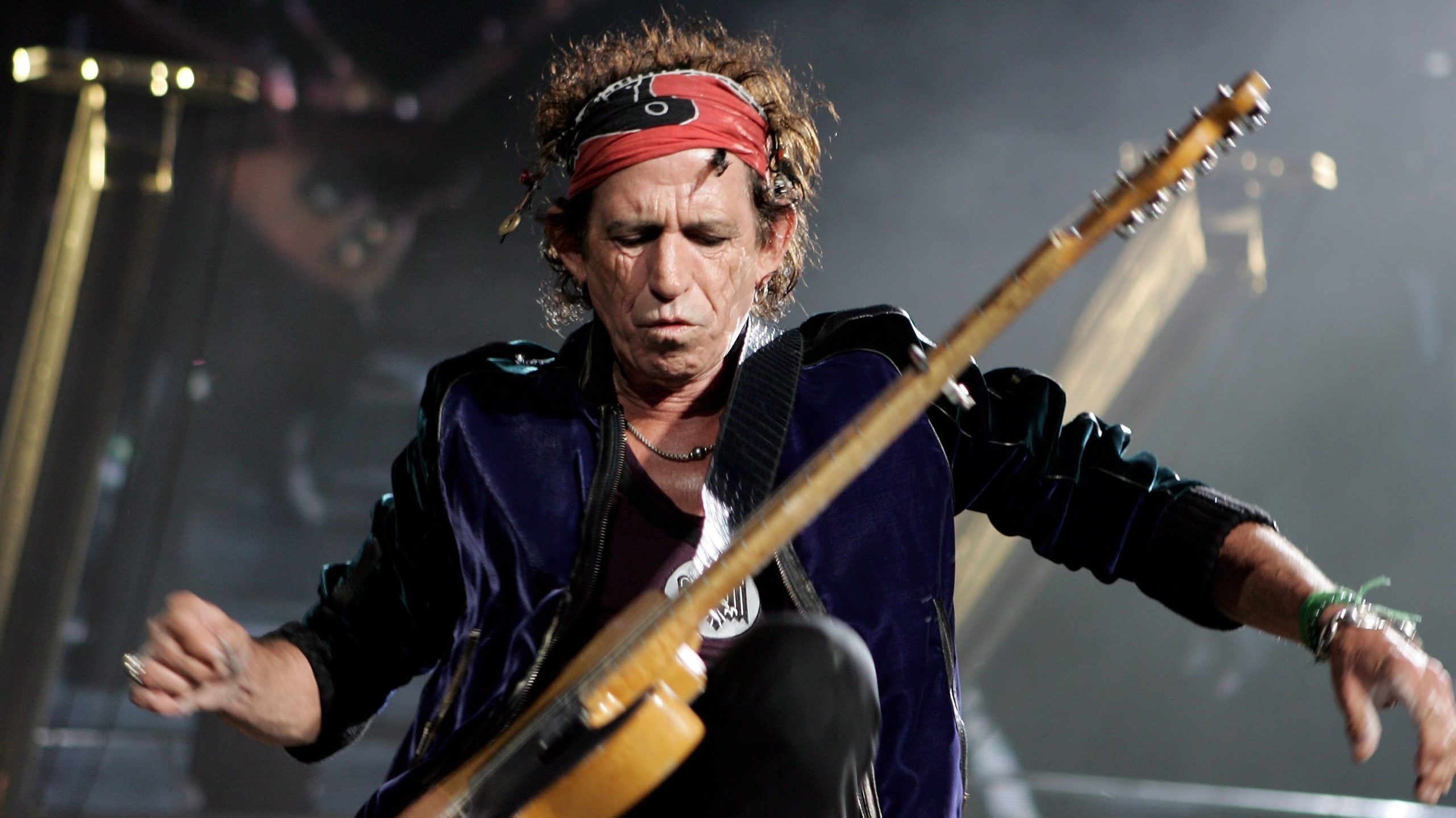 Keith Richards performing live on stage with intense focus, wearing a red bandana and dark velvet jacket while playing guitar.