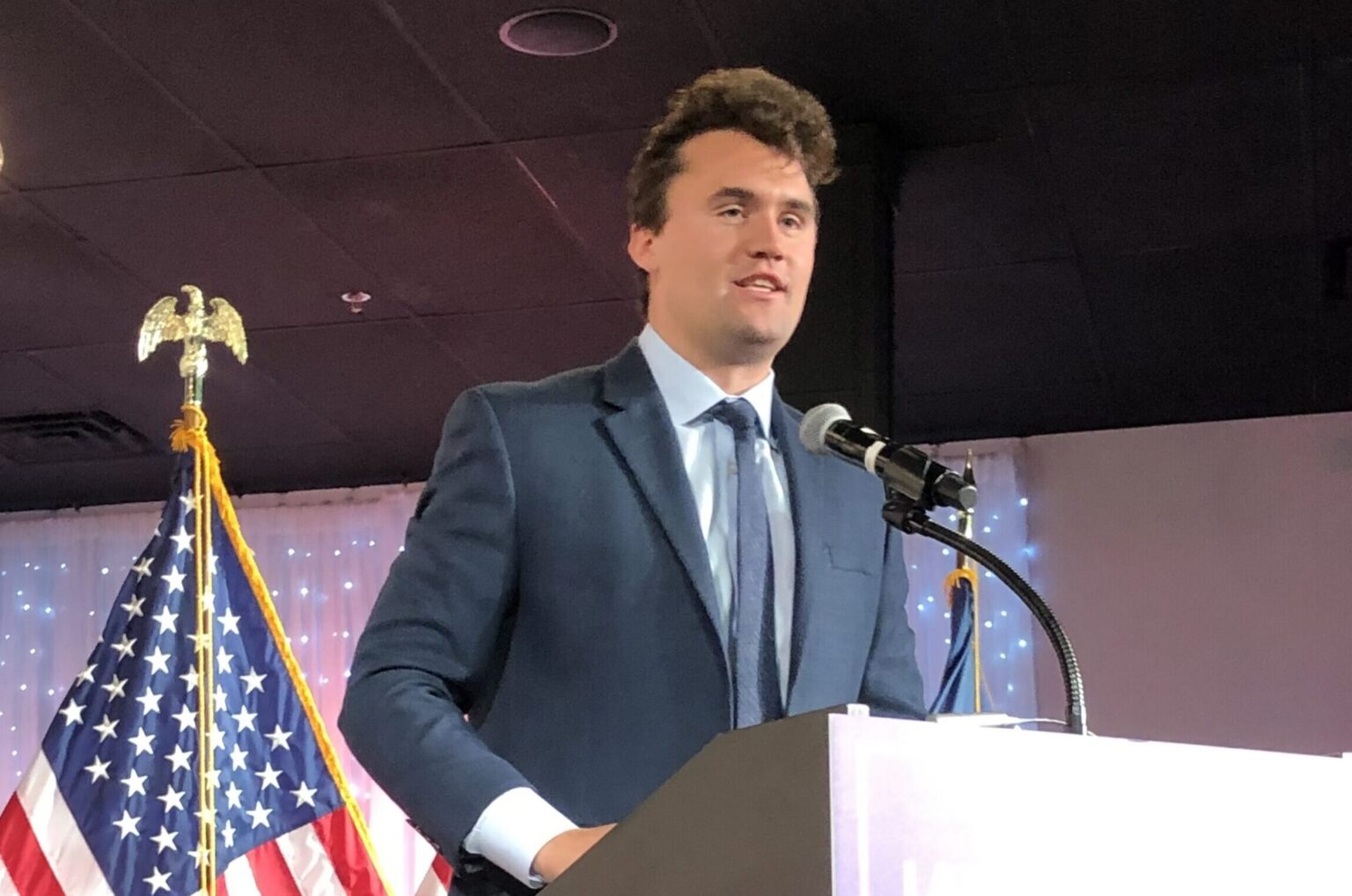 A younger Charlie Kirk passionately delivering a speech at a lectern, dressed in a suit and tie.
