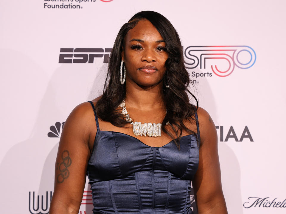 Claressa Shields wearing a blue dress and her "GWOAT" chain, smiling confidently at an event.