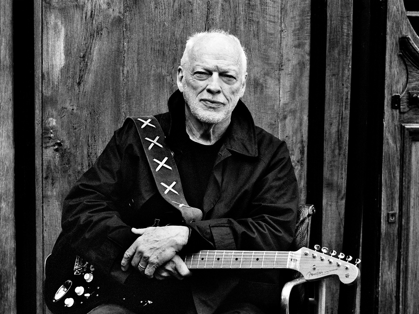 An older David Gilmour posing in a black and white portrait, holding his iconic Black Strat guitar with a calm, reflective expression.