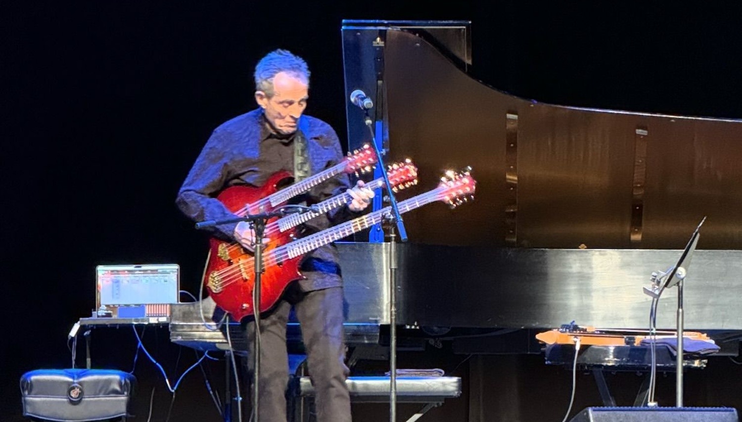An older John Paul Jones performing live on stage, playing his iconic triple-neck bass guitar under dramatic stage lighting.
