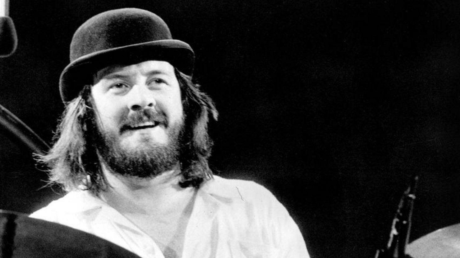 Black and white photo of John Bonham wearing a bowler hat while seated behind his drum kit.