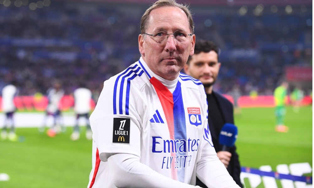 John Textor standing on the football pitch wearing an Olympique Lyonnais shirt, looking confident and engaged.