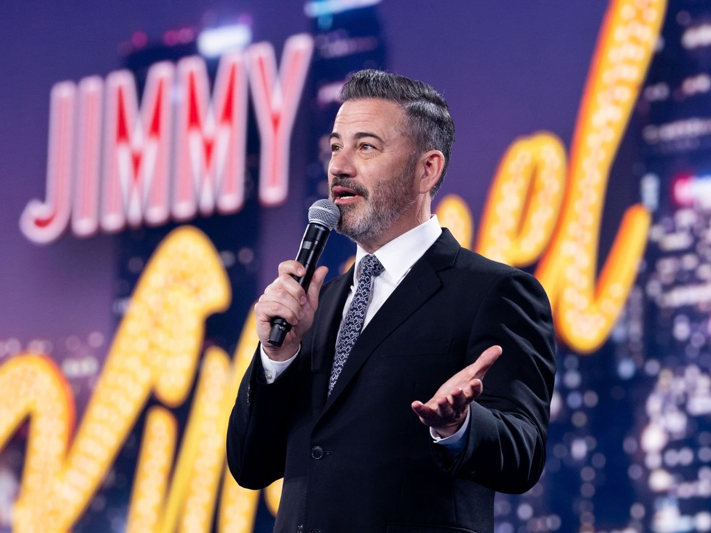 Jimmy Kimmel stands in front of the glowing neon sign for Jimmy Kimmel Live!, engaging with the audience during a live taping.