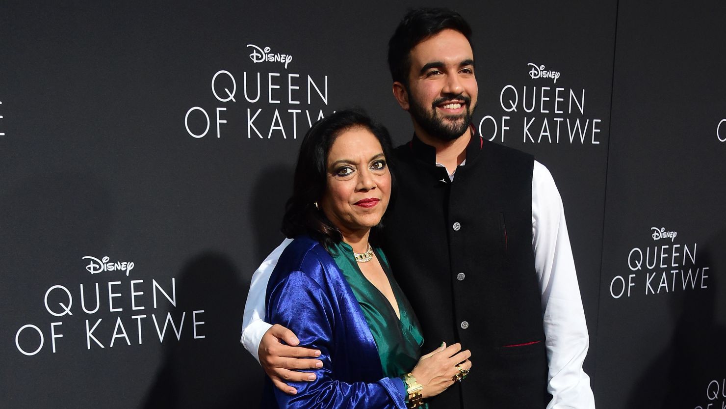 Zohran Mamdani stands beside his mother, filmmaker Mira Nair, on the red carpet, both dressed elegantly and smiling for the cameras.