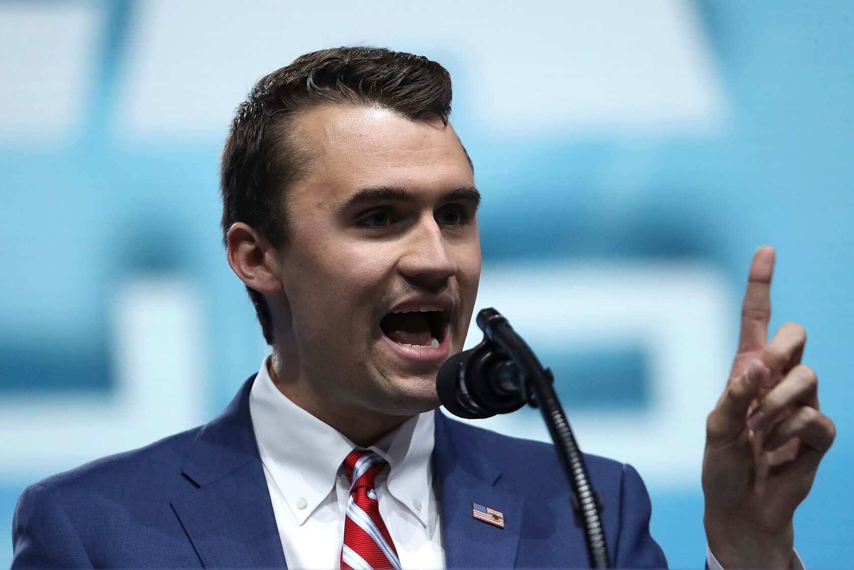 Charlie Kirk delivering a speech at a microphone, dressed in a sharp suit.