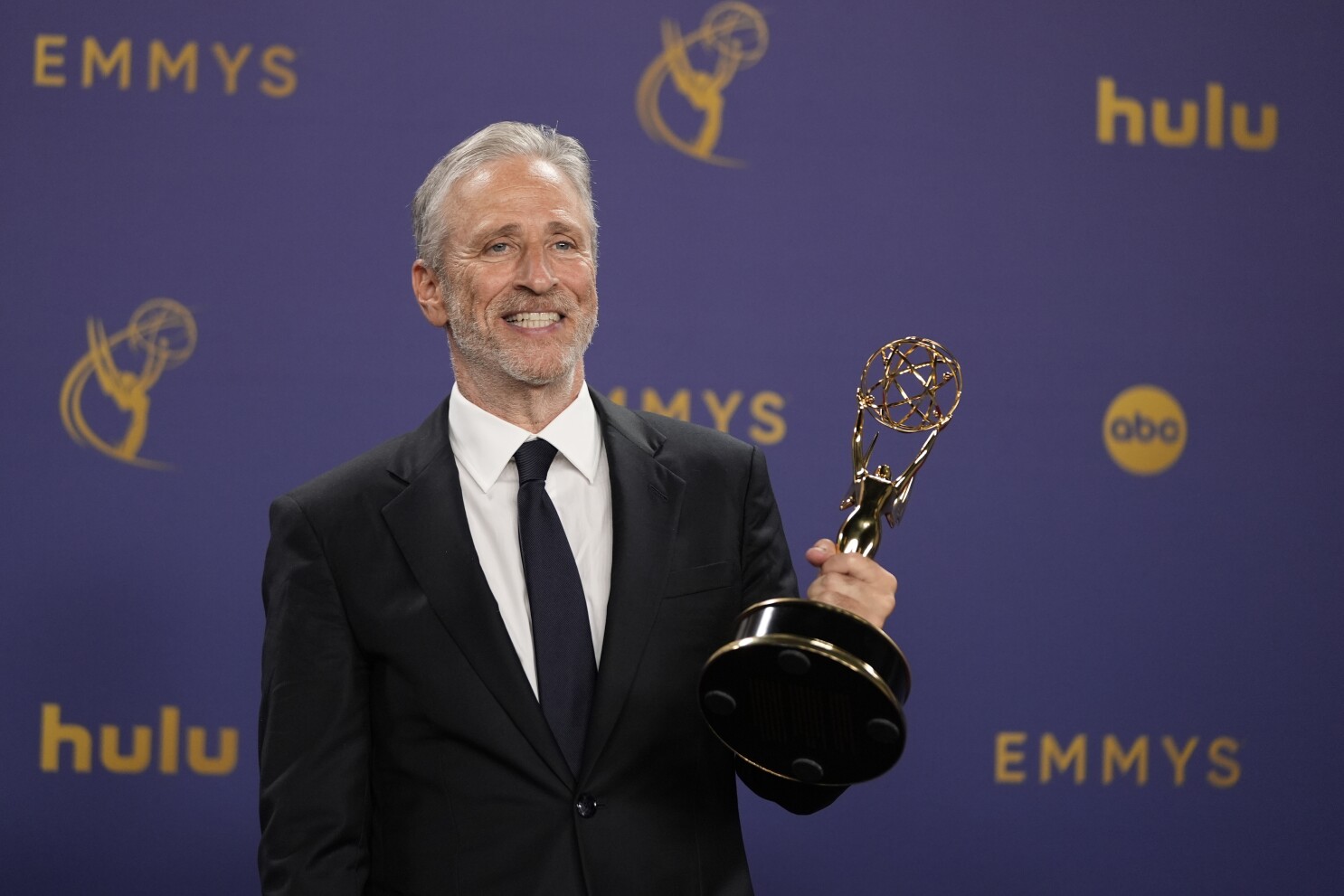 Jon Stewart holds his Emmy Award, smiling proudly in a classic suit after a major win