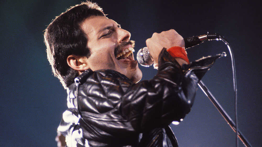 Freddie Mercury passionately singing into a microphone on stage, wearing a black leather jacket and a red wristband.