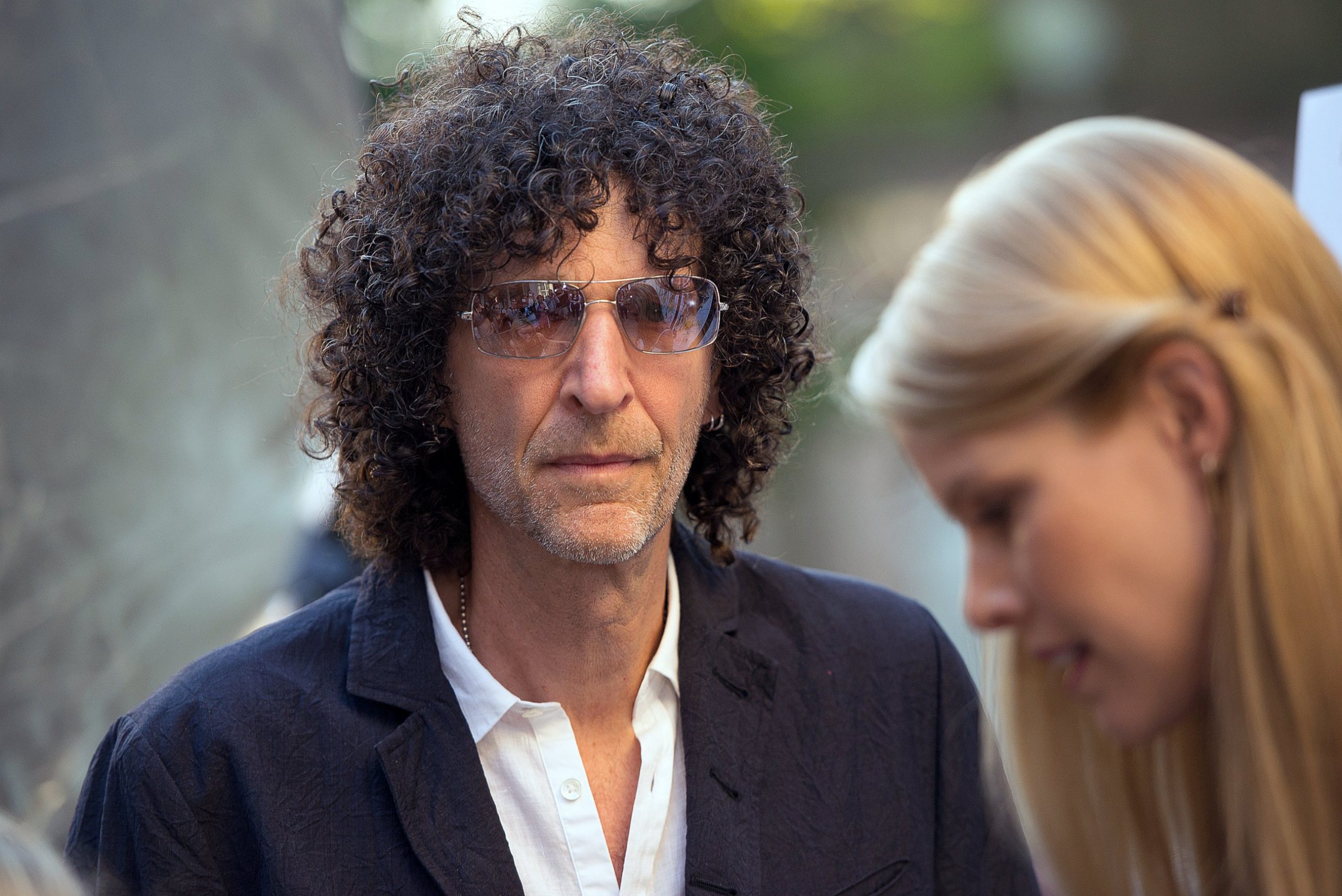 Howard Stern exits the studio wearing sunglasses and a stylish jacket, looking relaxed and confident as he steps outside.