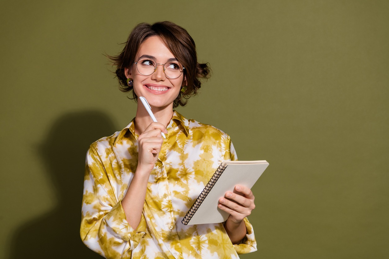 confident young businesswoman with thoughtful expression, holding pen and notebook, wearing stylish blouse, olive green backdrop