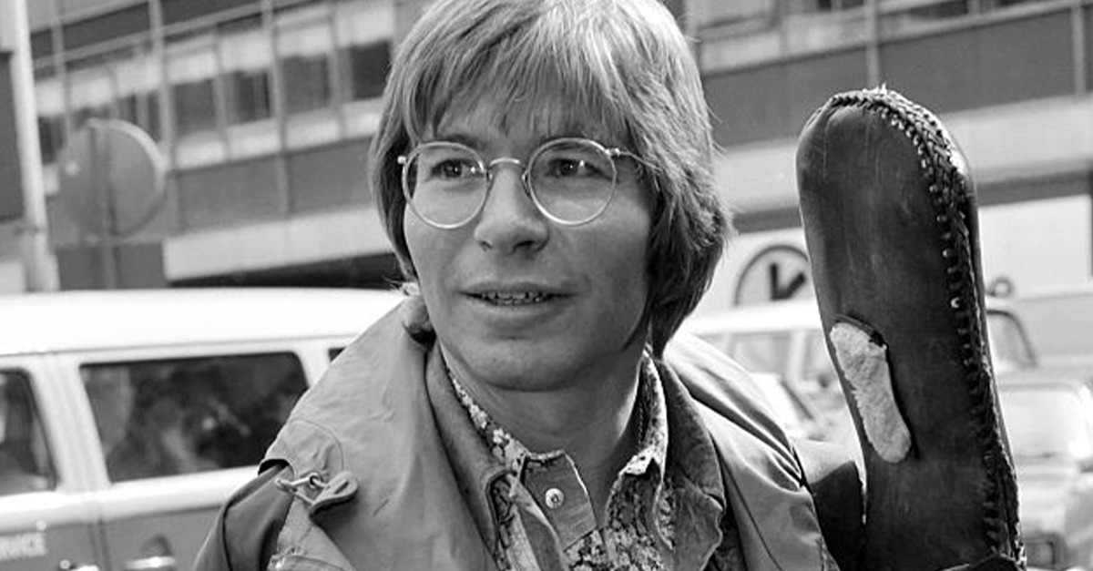Black and white photo of John Denver standing outdoors, holding a guitar case, wearing a casual jacket and looking thoughtfully into the distance.