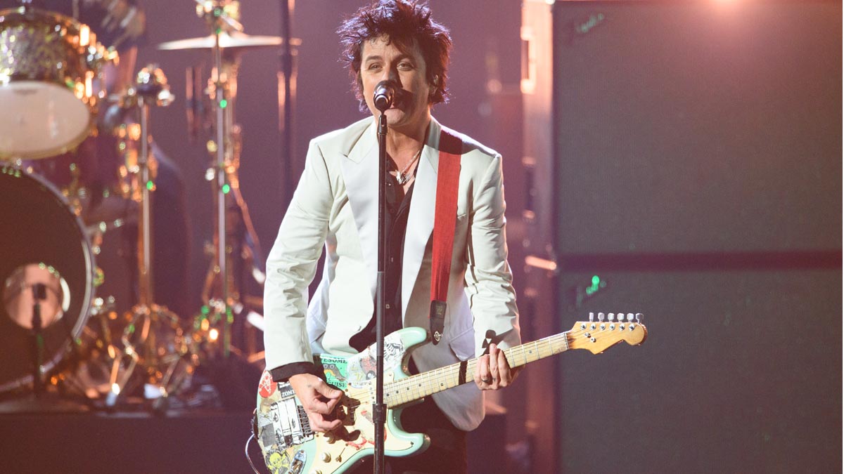 Billie Joe Armstrong performing on stage wearing a white blazer and playing a custom electric guitar, mid-performance under dramatic lighting.