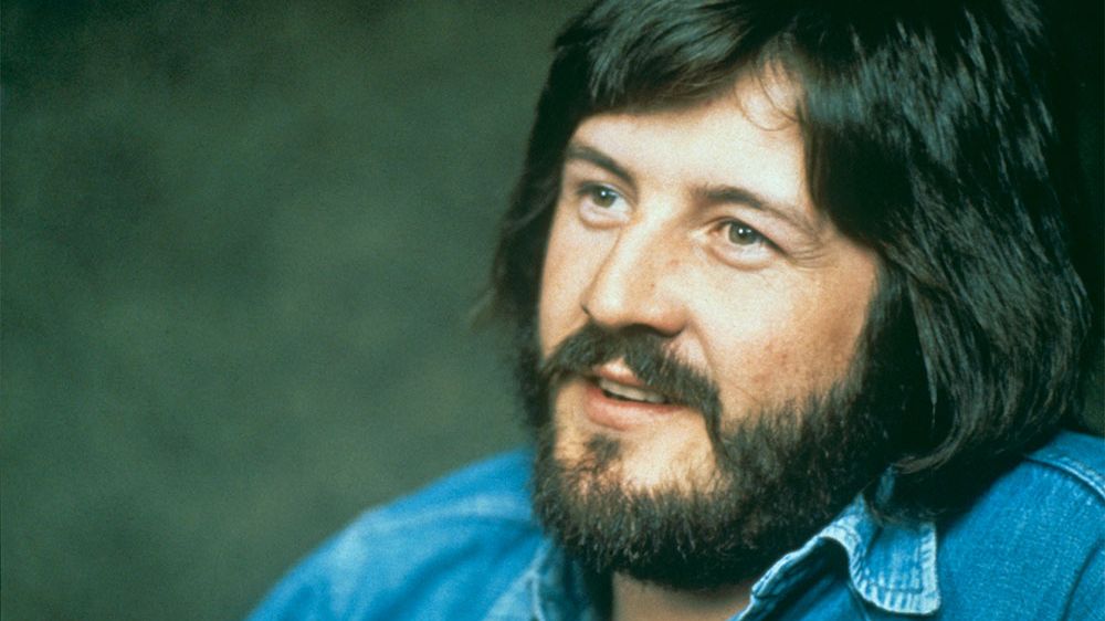 Portrait of John Bonham smiling warmly while wearing a blue denim shirt.