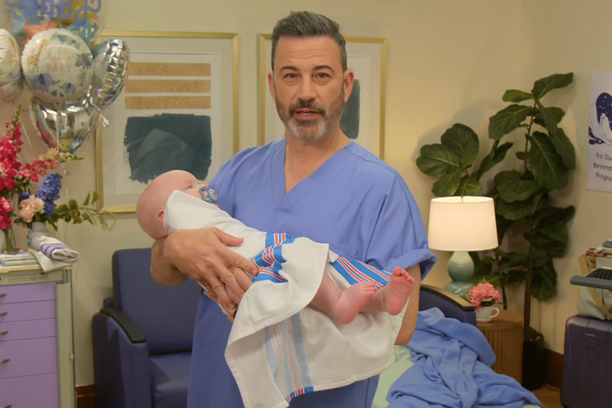 Jimmy Kimmel wearing hospital scrubs holds his grandchild tenderly, smiling down with pride and warmth.