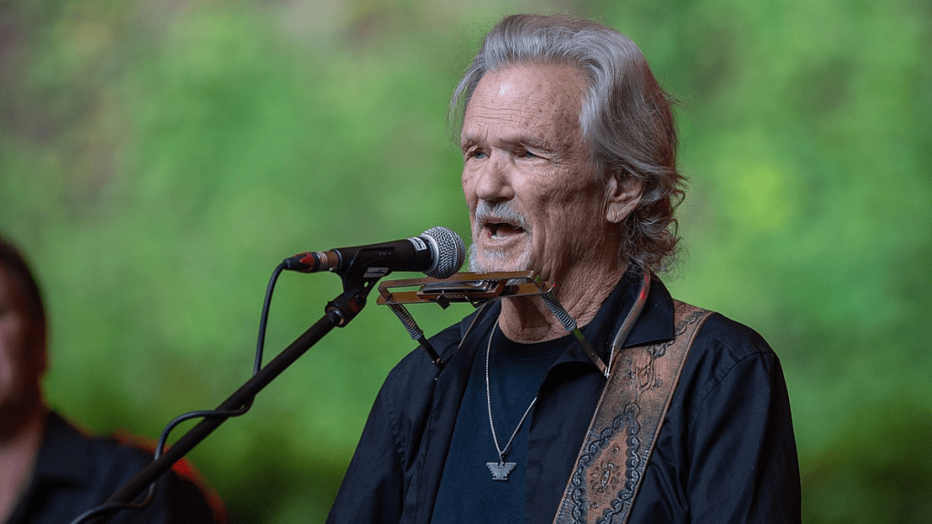 An elderly Kris Kristofferson performing outdoors on stage, holding a microphone and singing, surrounded by natural light and a casual audience setting.