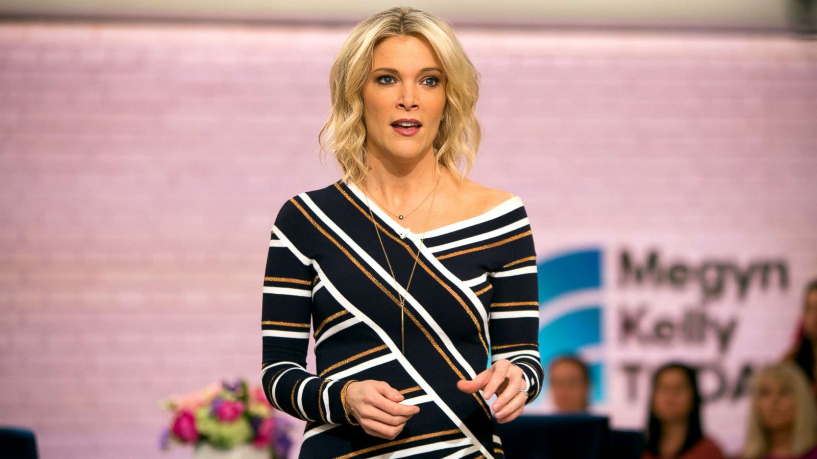 Megyn Kelly standing confidently on her show stage, addressing the live audience with emphasis.