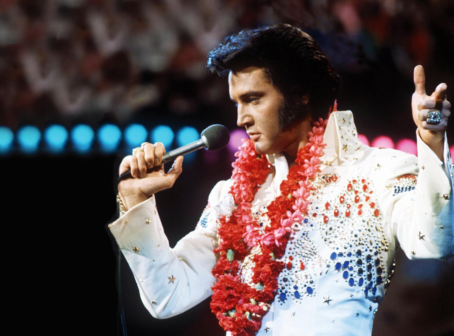 Elvis Presley performing on stage in a white jeweled jumpsuit with red and pink flower leis, holding a microphone and gesturing with his hand.