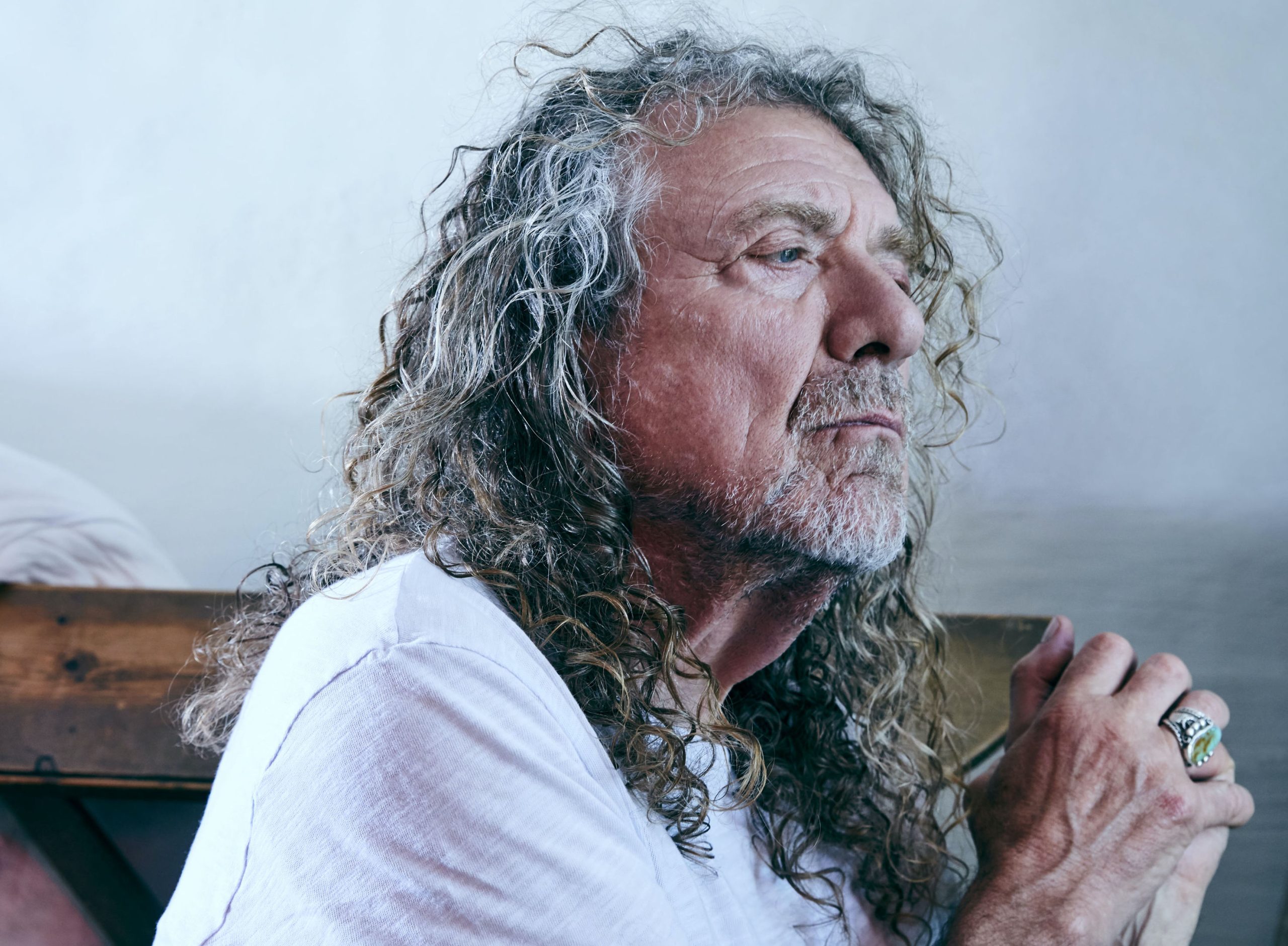 Robert Plant in 2025, deep in thought, with long curly hair and a white shirt, gazing contemplatively to the side.