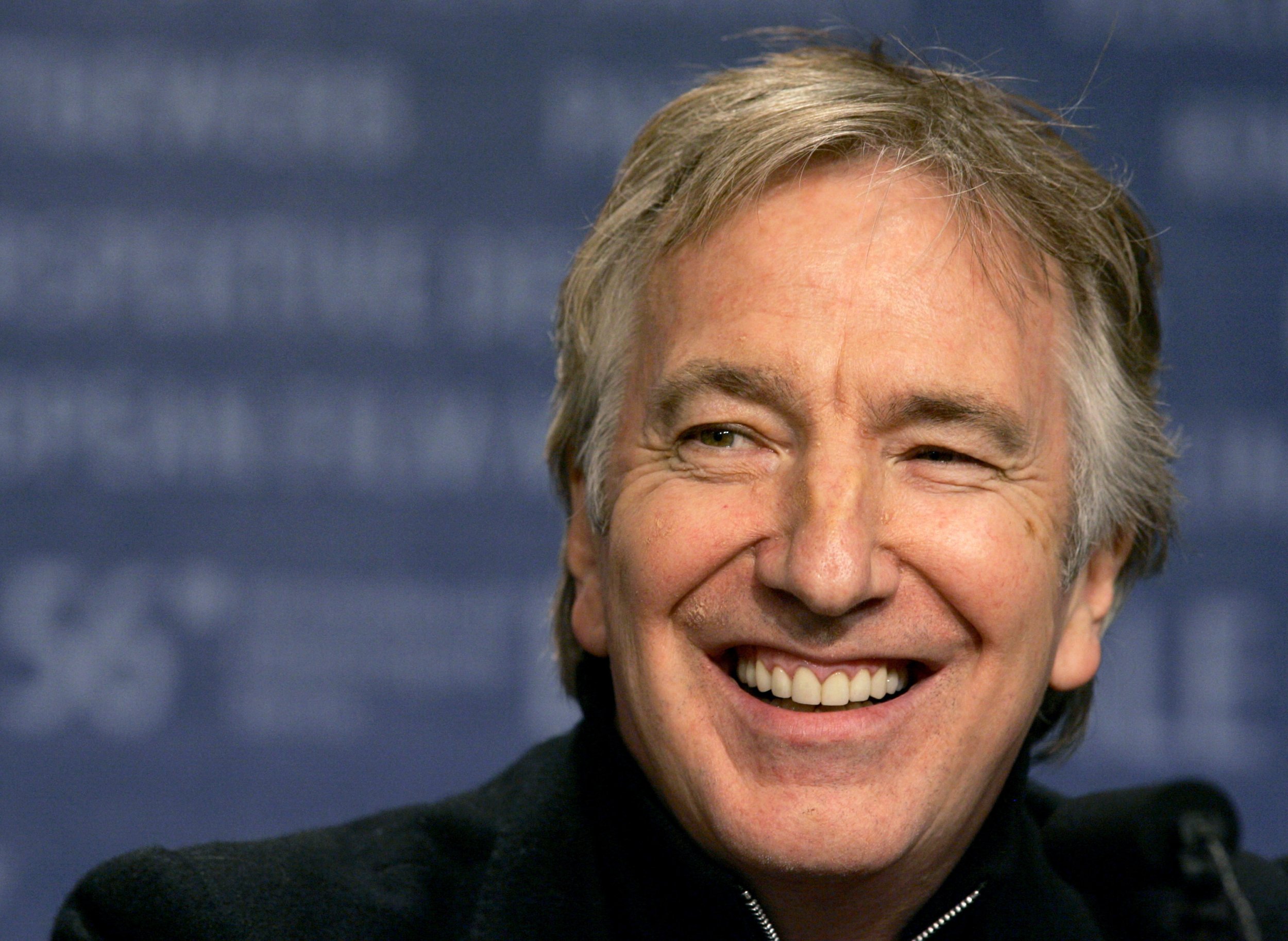 Alan Rickman smiling warmly in a candid moment, dressed in a dark jacket.