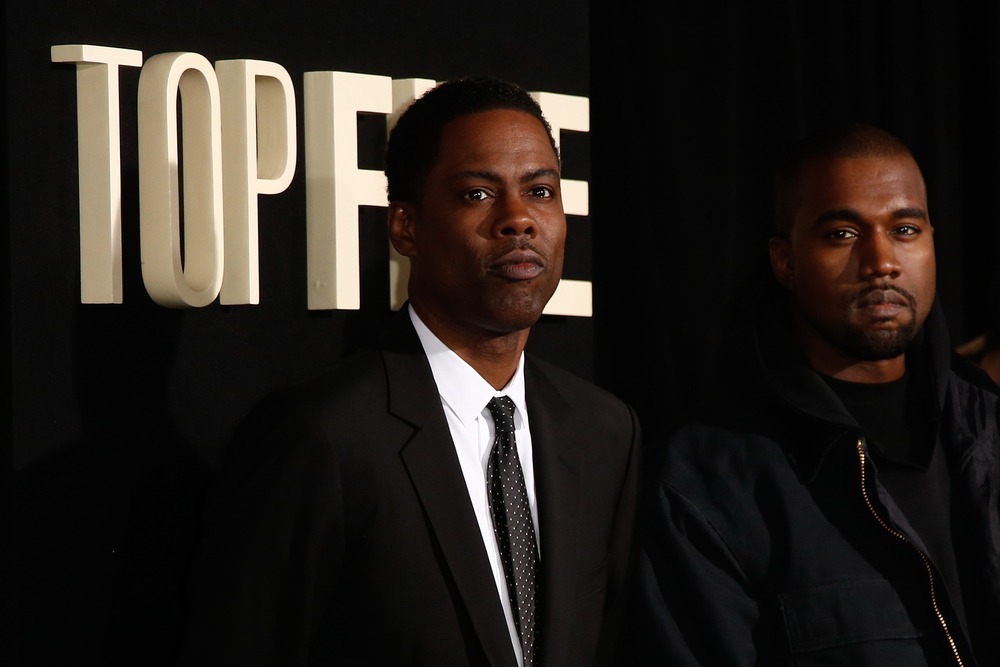 Chris Rock looking serious while standing next to Kanye West on the red carpet, both dressed in stylish attire.