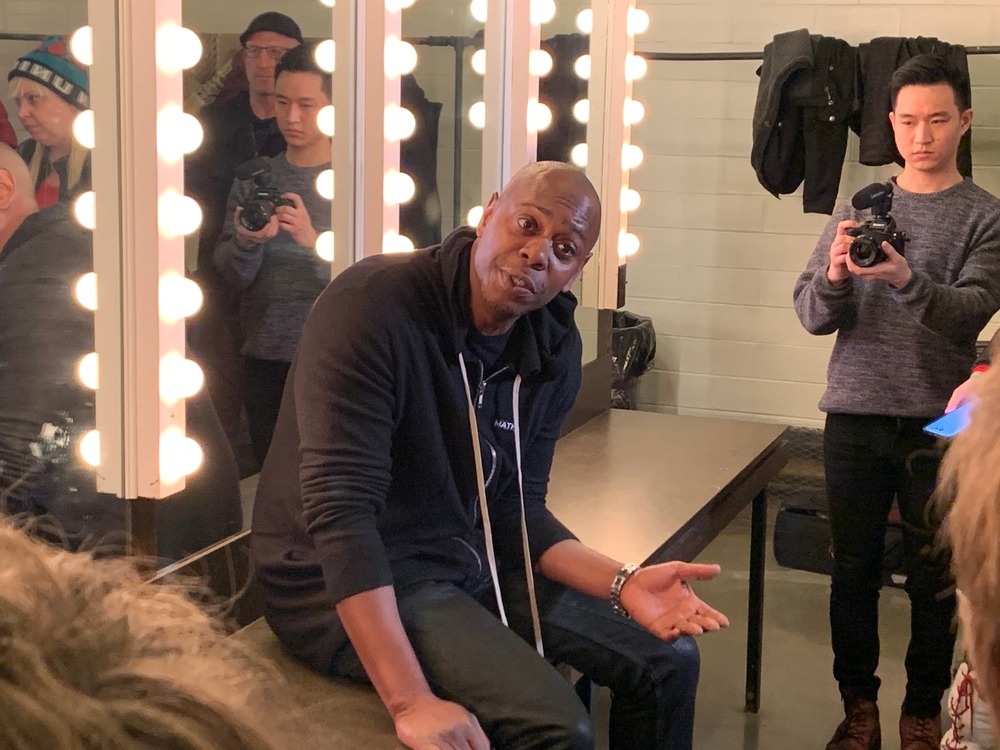 Dave Chappelle sits in his dressing room, surrounded by glowing mirror lights, as he gives a candid interview before taking the stage.