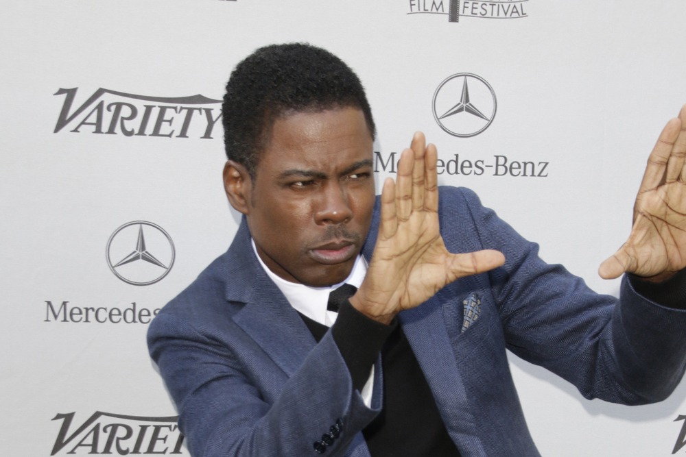 Chris Rock striking a confident pose on the red carpet with both hands raised like a director, wearing a sharp suit.