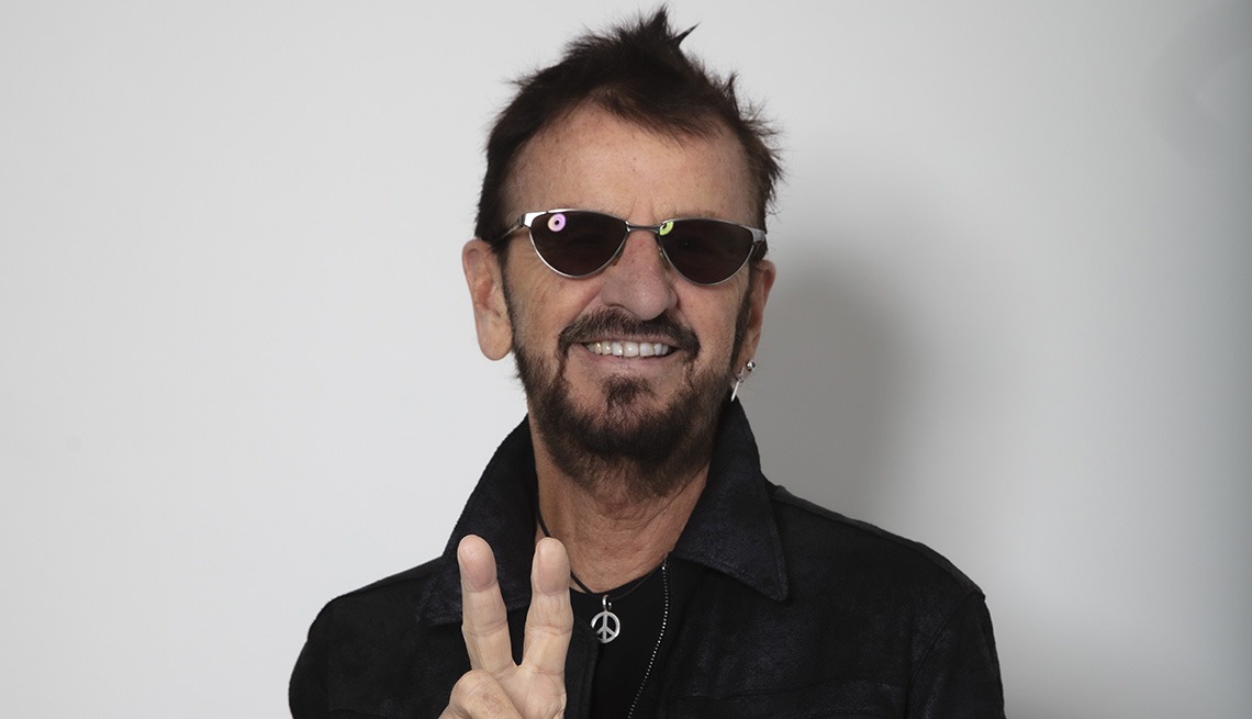Ringo Starr smiling while giving a peace sign, wearing dark sunglasses and a black jacket, against a plain white background.