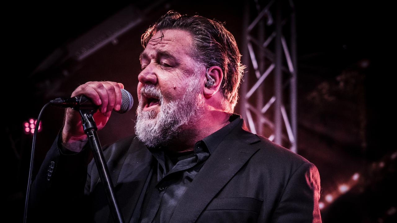 Russell Crowe passionately singing on stage with his band, holding a microphone.