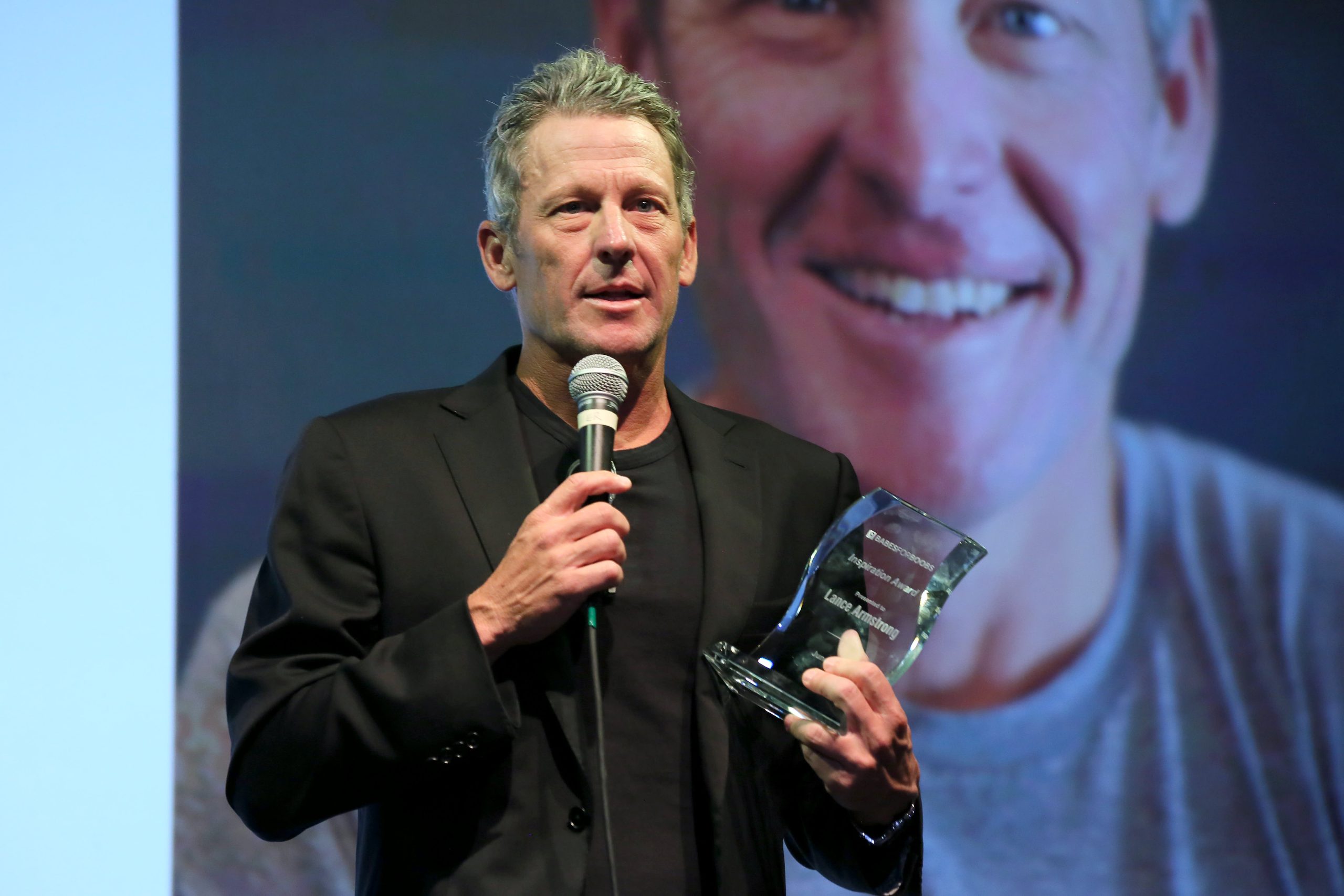 Lance Armstrong speaking passionately at a corporate event, dressed in a dark blazer and holding a microphone.