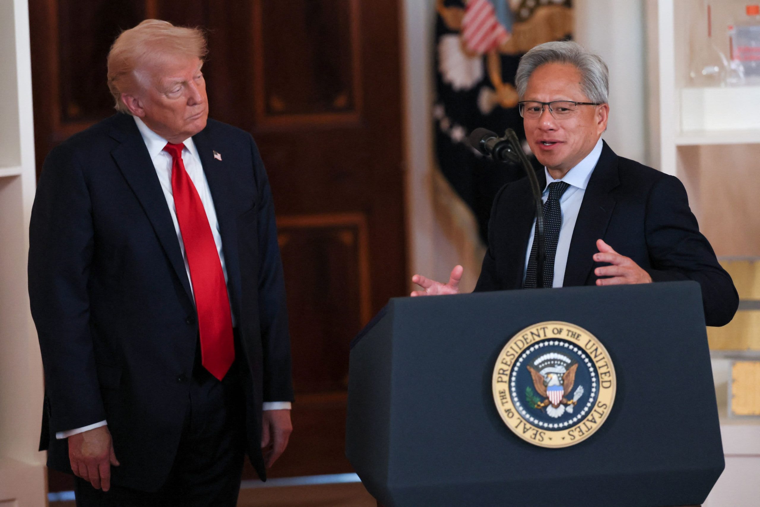Donald Trump listens as Nvidia CEO Jensen Huang speaks at a podium with the presidential seal during an AI summit at the White House, July 2025.